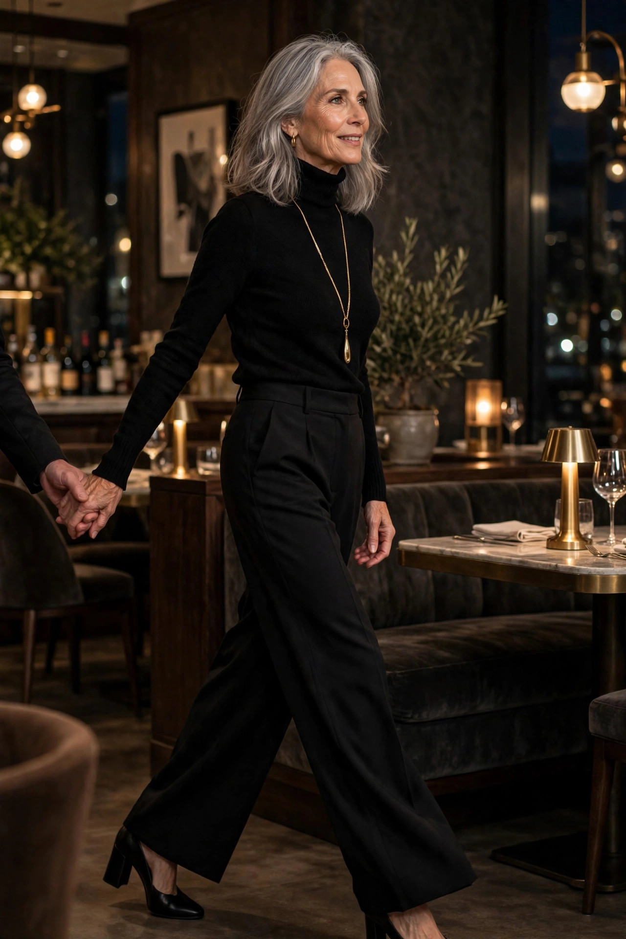 Woman in her 50s in black turtleneck and wide-leg trousers with long gold pendant in upscale restaurant